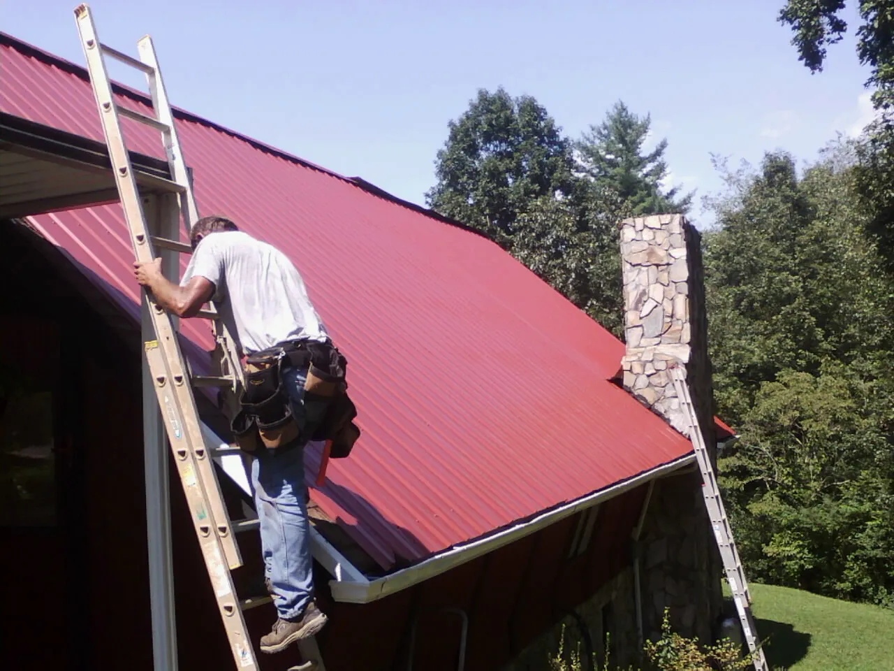 Roofer on a ladder beside a red metal roof and stone chimney — Soffit & Fascia Repair by Roofing Brothers in Palatka